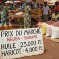 Des marchandes et des sacs de grains colorés sous des parasols dans un marché en plein air, avec un panneau au premier plan indiquant les prix de l'huile et des haricots à Bukavu.