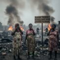 Trois femmes enceintes se tiennent debout au premier plan, couvrant leur bouche et leur nez, devant des ruines fumantes et un panneau en bois portant l'inscription « BUKAVU : INCENDIES RÉPÉRTÉS, SANTÉ FEMMES ENCEINTES EN DANGER », tandis que des pompiers luttent contre les flammes en arrière-plan.