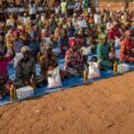 Un grand groupe de femmes assises par terre en extérieur tient des sacs de provisions et des bouteilles d'huile devant une grande tente blanche.