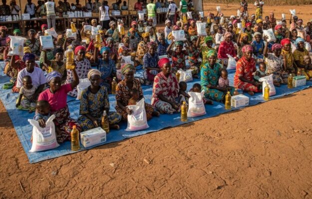 Un grand groupe de femmes assises par terre en extérieur tient des sacs de provisions et des bouteilles d'huile devant une grande tente blanche.