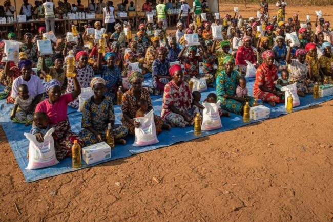 Un grand groupe de femmes assises par terre en extérieur tient des sacs de provisions et des bouteilles d'huile devant une grande tente blanche.