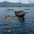 Une épave de bateau en bois flotte à demi-submergée sur le lac Kivu avec des débris à la surface, tandis que de petites pirogues naviguent au loin.