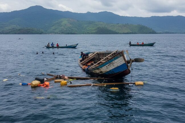 Une épave de bateau en bois flotte à demi-submergée sur le lac Kivu avec des débris à la surface, tandis que de petites pirogues naviguent au loin.